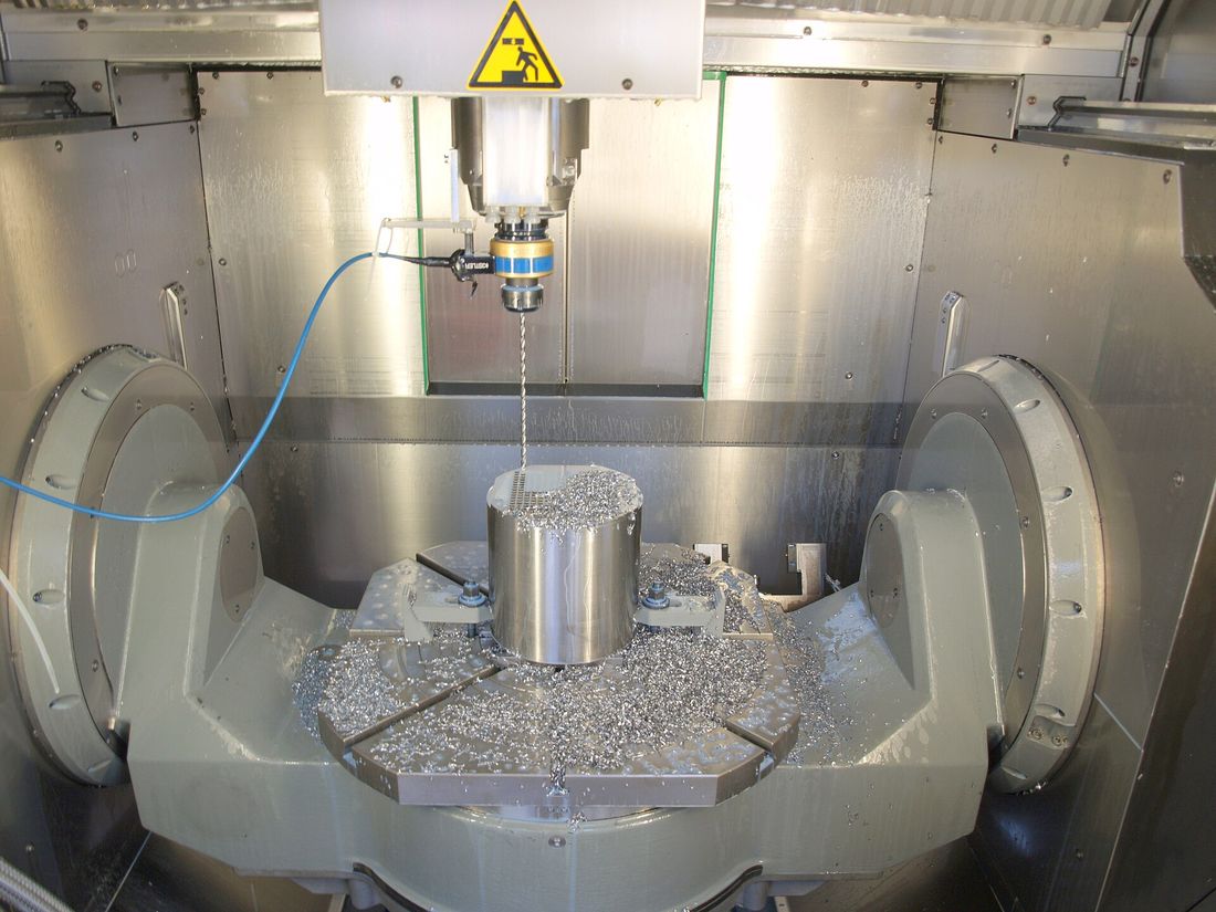Cylindrical stainless part being drilled on a five-axis trunnion machining center, surrounded by chips